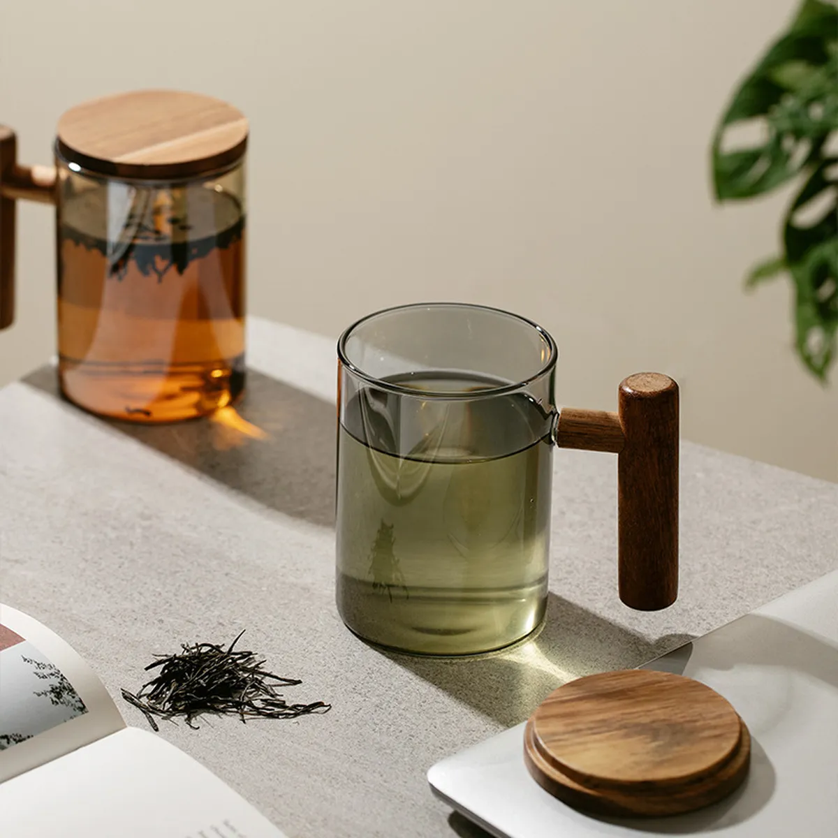 Amber and tinted grey glass mugs with wooden lids, loose tea leaves beside them